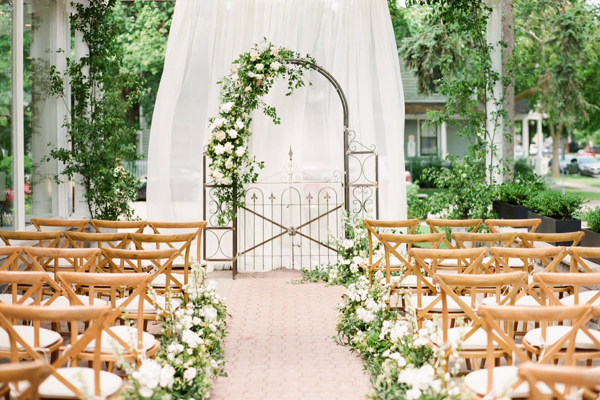 Outdoor wedding ceremony setup featuring wooden chairs aligned in rows on both sides of a flower-lined aisle. A white curtain backdrop and a metal arch adorned with greenery and white flowers are at the front, with trees visible in the background. 124 on Queen Hotel and Spa | Hotel & Spa Niagara-on-the-lake | 1 (855) 988-4552