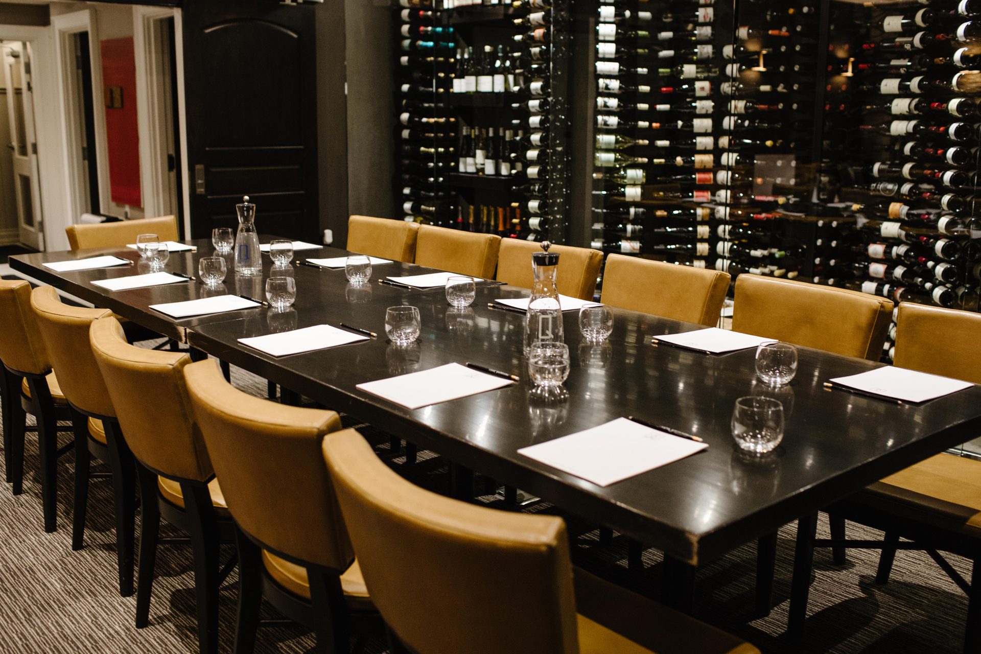 A long black table is set for a meeting or dinner with beige chairs around it. Empty glasses, water carafes, and papers are placed on the table. The background features wine racks filled with bottles, creating a sophisticated atmosphere. 124 on Queen Hotel and Spa | Hotel & Spa Niagara-on-the-lake | 1 (855) 988-4552