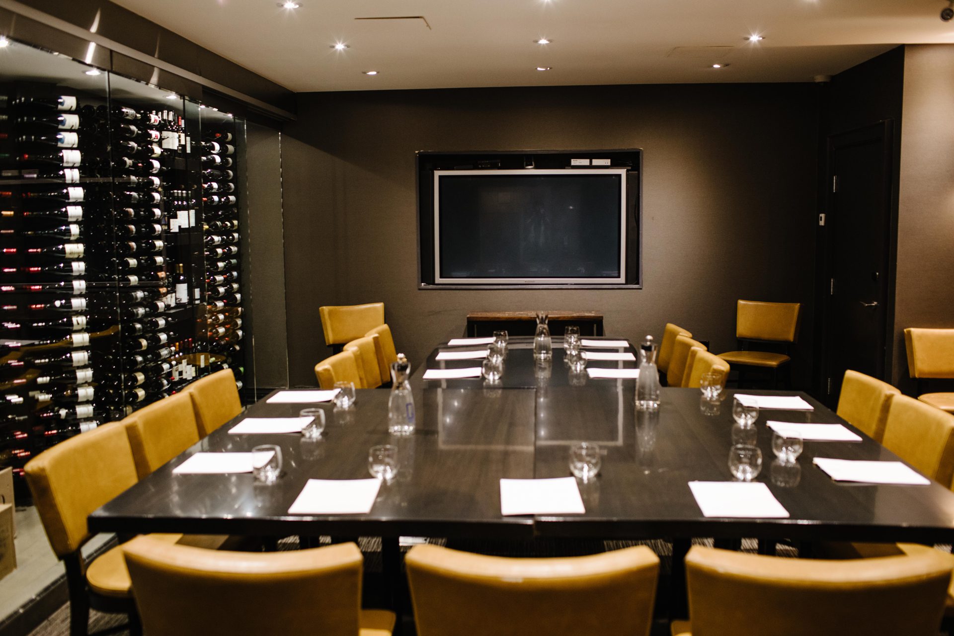 A modern conference room with a rectangular table surrounded by tan chairs. The table is set with notepads and glasses. A large screen is mounted on a dark wall, and a wine rack is visible to the left. The room is well-lit with recessed lighting. 124 on Queen Hotel and Spa | Hotel & Spa Niagara-on-the-lake | 1 (855) 988-4552