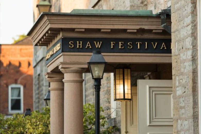 Entrance to the Shaw Festival building, featuring a classical design with columns and a black sign with gold lettering. The open door and lantern-style light provide a welcoming atmosphere. Brick buildings are visible in the background. 124 on Queen Hotel and Spa | Hotel & Spa Niagara-on-the-lake | 1 (855) 988-4552