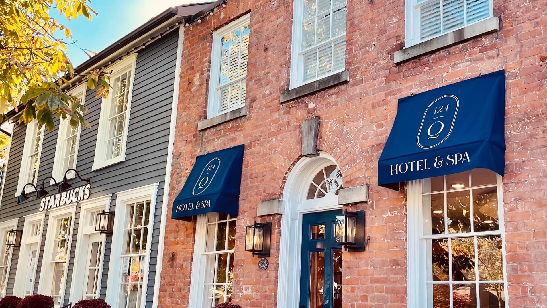 A charming street view of a red brick building with two blue awnings labeled 124 Hotel & Spa next to a gray building with a Starbucks sign. Bright windows reflect a sunny sky, and autumn leaves are partially visible on the left. 124 on Queen Hotel and Spa | Hotel & Spa Niagara-on-the-lake | 1 (855) 988-4552