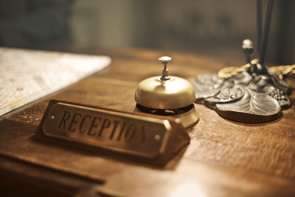 Close-up of a wooden reception desk featuring a brass bell and a sign labeled RECEPTION. A decorative item with leaf patterns is partially visible nearby, set against a softly lit background. 124 on Queen Hotel and Spa | Hotel & Spa Niagara-on-the-lake | 1 (855) 988-4552 Close-up of a wooden reception desk featuring a brass bell and a sign labeled RECEPTION. A decorative item with leaf patterns is partially visible nearby, set against a softly lit background. 124 on Queen Hotel and Spa | Hotel & Spa Niagara-on-the-lake | 1 (855) 988-4552