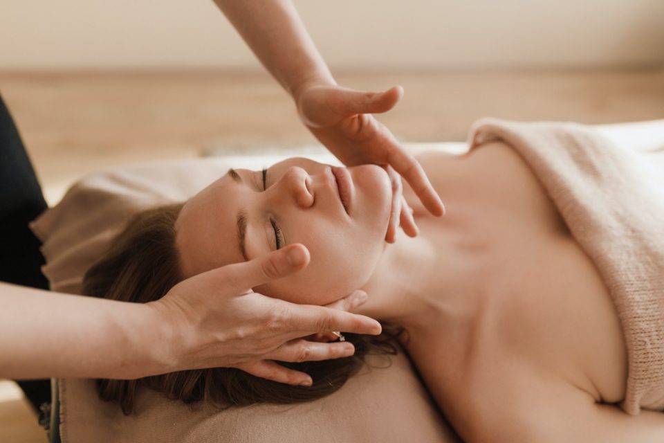 A person receiving a facial massage while lying down, covered with a towel. Hands gently massage their face, creating a calm and relaxing atmosphere. The setting appears serene, with soft lighting. 124 on Queen Hotel and Spa | Hotel & Spa Niagara-on-the-lake | 1 (855) 988-4552