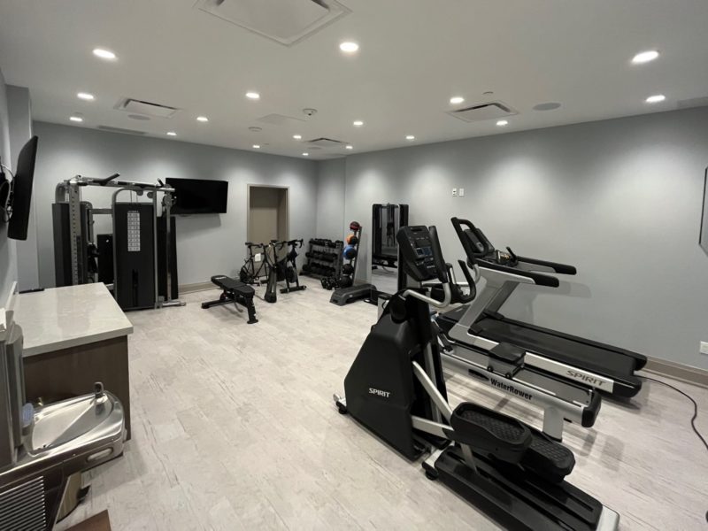 A modern gym with exercise equipment including treadmills, stationary bikes, and strength machines. The room features light gray walls, overhead lighting, and a wall-mounted TV. A drinking fountain and weights are also visible. 124 on Queen Hotel and Spa | Hotel & Spa Niagara-on-the-lake | 1 (855) 988-4552