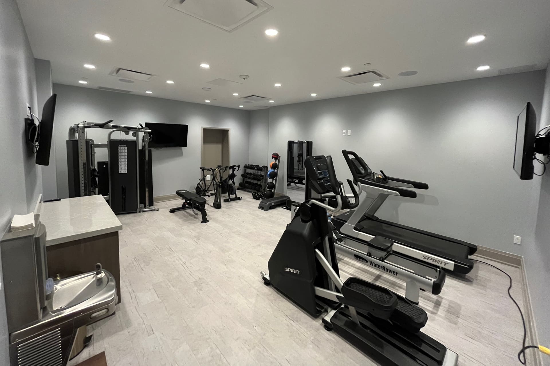 A modern gym with exercise equipment including treadmills, stationary bikes, and strength machines. The room features light gray walls, overhead lighting, and a wall-mounted TV. A drinking fountain and weights are also visible. 124 on Queen Hotel and Spa | Hotel & Spa Niagara-on-the-lake | 1 (855) 988-4552 A modern gym with exercise equipment including treadmills, stationary bikes, and strength machines. The room features light gray walls, overhead lighting, and a wall-mounted TV. A drinking fountain and weights are also visible. 124 on Queen Hotel and Spa | Hotel & Spa Niagara-on-the-lake | 1 (855) 988-4552