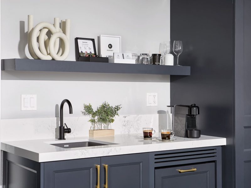 Modern kitchen area with a black sink and faucet, dark cabinets with gold handles, and a white countertop. A shelf above holds decor, glassware, and a coffee machine with two espresso cups. A small plant is placed near the sink. 124 on Queen Hotel and Spa | Hotel & Spa Niagara-on-the-lake | 1 (855) 988-4552