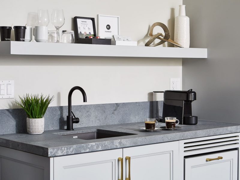 Modern kitchen with a dark gray countertop, black faucet, and a coffee machine. Two cups of espresso sit on the counter. A white floating shelf above holds decor items, coffee cups, and glasses. A small potted plant is near the sink. 124 on Queen Hotel and Spa | Hotel & Spa Niagara-on-the-lake | 1 (855) 988-4552