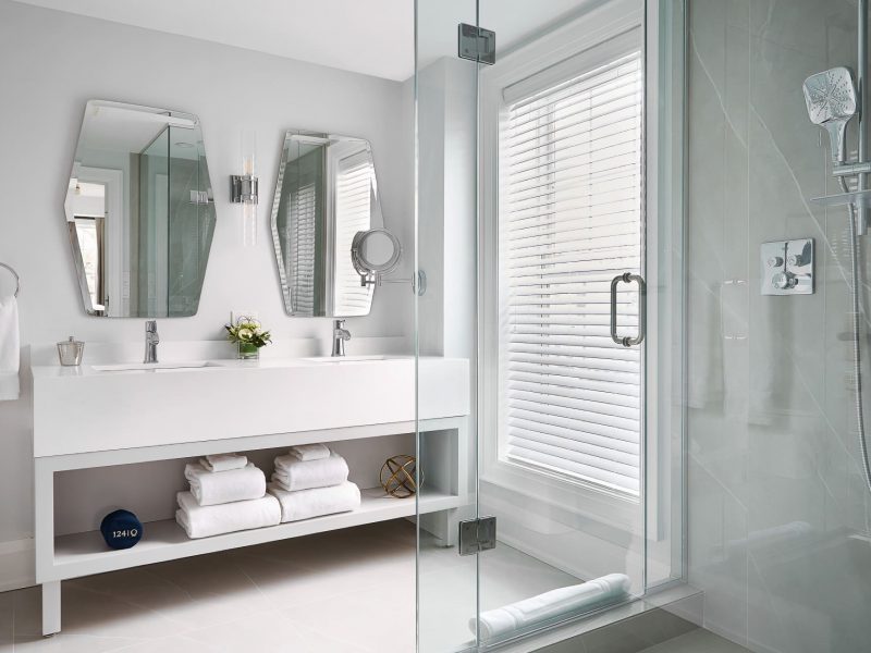 Modern bathroom with glass-enclosed shower, dual sinks on a white vanity, two geometric mirrors, and a window with blinds. White towels are neatly stacked below the vanity; decor includes a small plant and wall-mounted light fixtures. 124 on Queen Hotel and Spa | Hotel & Spa Niagara-on-the-lake | 1 (855) 988-4552