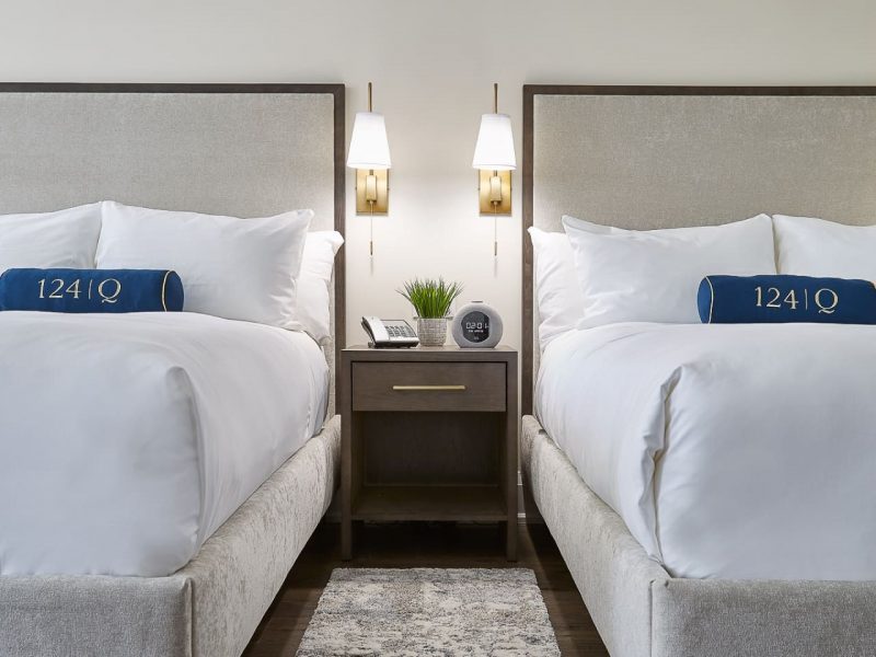 A hotel room with two neatly made double beds, each with white linens and blue pillows labeled 1241. A wooden nightstand with a clock, phone, potted plant, and wall-mounted lamps is between the beds. A rug lies on the wooden floor. 124 on Queen Hotel and Spa | Hotel & Spa Niagara-on-the-lake | 1 (855) 988-4552