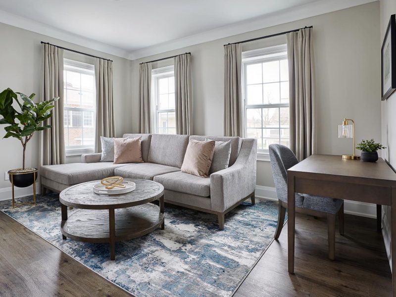A modern living room with a gray sectional sofa and two pink pillows, a round coffee table, a blue and gray rug, and a wooden desk with a chair. There is a potted plant in the corner and three tall windows with beige curtains. 124 on Queen Hotel and Spa | Hotel & Spa Niagara-on-the-lake | 1 (855) 988-4552