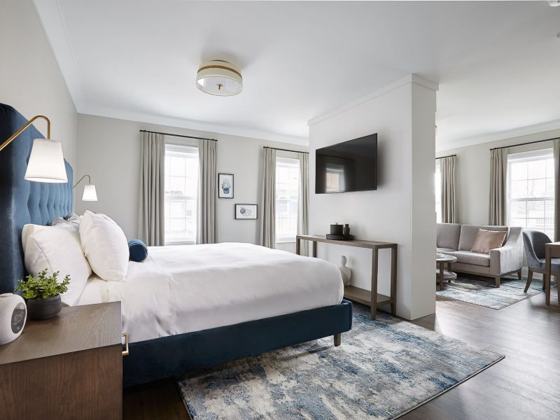 A bright, modern hotel room with a large bed, white linens, and a blue headboard. A wall-mounted TV is above a minimalist console table. Theres a seating area with a sofa and chairs. Multiple windows let in natural light, and decorative art is on the walls. 124 on Queen Hotel and Spa | Hotel & Spa Niagara-on-the-lake | 1 (855) 988-4552