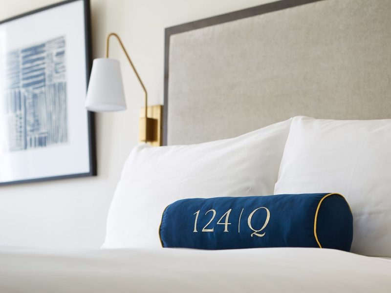A neatly made hotel bed with a blue cylindrical pillow featuring 124 | Q in white. A soft gray headboard and a framed abstract artwork adorn the wall along with a modern wall-mounted lamp. 124 on Queen Hotel and Spa | Hotel & Spa Niagara-on-the-lake | 1 (855) 988-4552