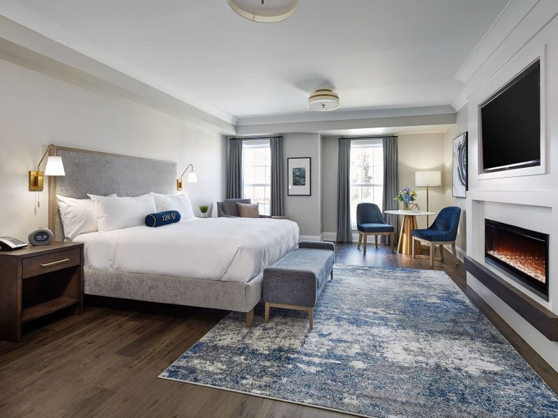 A spacious hotel room with a large bed, bedside tables, and a TV mounted above a modern fireplace. Two chairs and a small table are by the window. The room features light walls, hardwood flooring, and a patterned blue rug. 124 on Queen Hotel and Spa | Hotel & Spa Niagara-on-the-lake | 1 (855) 988-4552