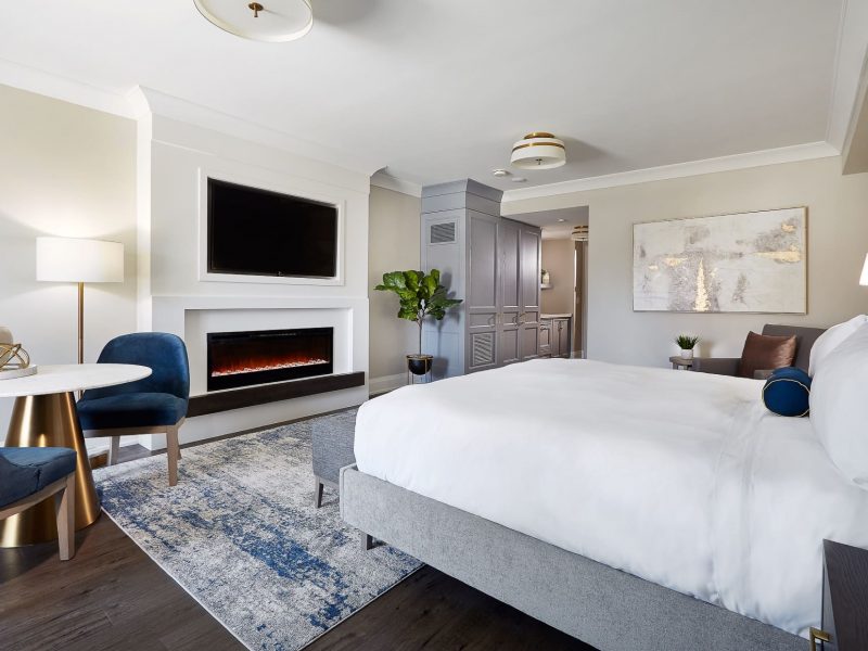 A modern hotel room with a large bed, blue and white decor, and a cozy sitting area. Theres a wall-mounted TV above a sleek electric fireplace, abstract artwork on the wall, and a large leafy plant. 124 on Queen Hotel and Spa | Hotel & Spa Niagara-on-the-lake | 1 (855) 988-4552