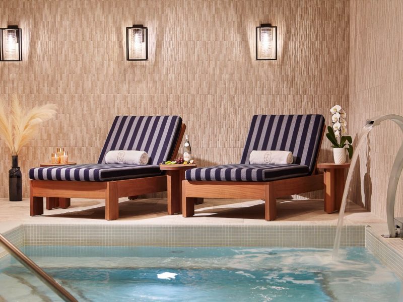 Two striped lounge chairs with rolled towels sit beside a small indoor pool. A vase of pampas grass and a potted orchid decorate the area. Water flows into the pool, and warm lighting creates a serene atmosphere. 124 on Queen Hotel and Spa | Hotel & Spa Niagara-on-the-lake | 1 (855) 988-4552