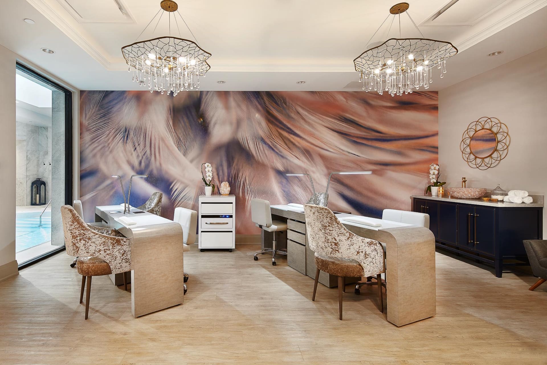 Luxurious nail salon interior with plush chairs, sleek white nail stations, and a large abstract mural wall. Elegant chandeliers hang from the ceiling. A large window shows a pool area, and a sideboard accessorized with decor accents is visible. 124 on Queen Hotel and Spa | Hotel & Spa Niagara-on-the-lake | 1 (855) 988-4552