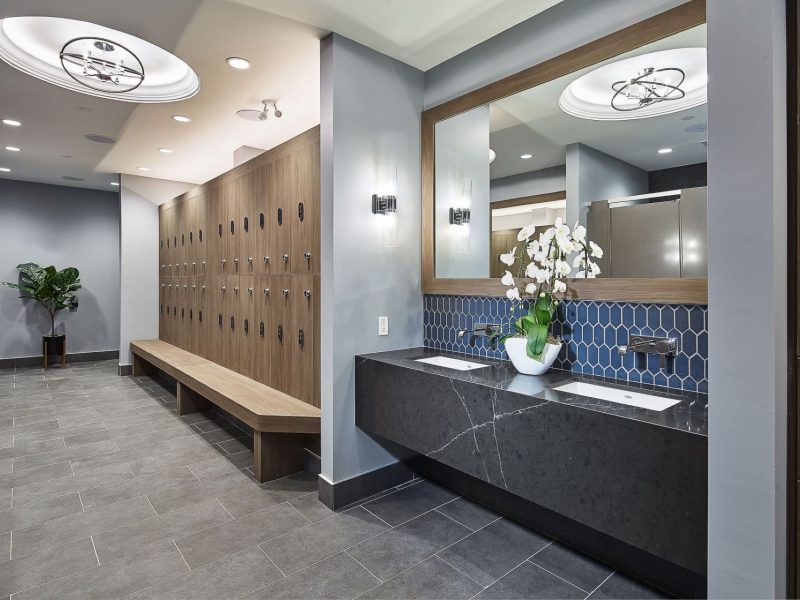 Modern locker room with gray tile flooring, a long wooden bench, and rows of lockers on the left. To the right, a countertop with two sinks and a large mirror above. A potted plant and stylish ceiling lights enhance the rooms aesthetics. 124 on Queen Hotel and Spa | Hotel & Spa Niagara-on-the-lake | 1 (855) 988-4552