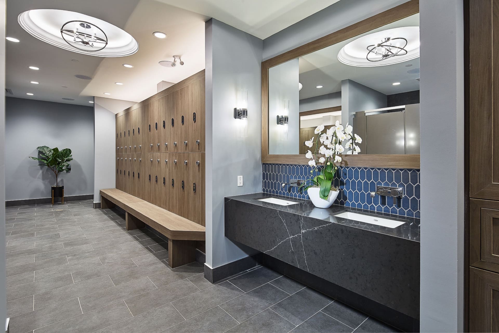 Modern locker room with gray tile flooring, a long wooden bench, and rows of lockers on the left. To the right, a countertop with two sinks and a large mirror above. A potted plant and stylish ceiling lights enhance the rooms aesthetics. 124 on Queen Hotel and Spa | Hotel & Spa Niagara-on-the-lake | 1 (855) 988-4552