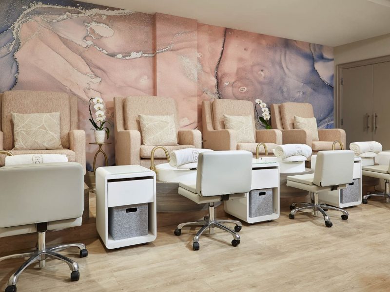 A spa with plush beige chairs and white footstools arranged for pedicure services. Each chair has a pillow and rolled towel. The background features a marbled pink and gray mural. The floor is light wood. 124 on Queen Hotel and Spa | Hotel & Spa Niagara-on-the-lake | 1 (855) 988-4552