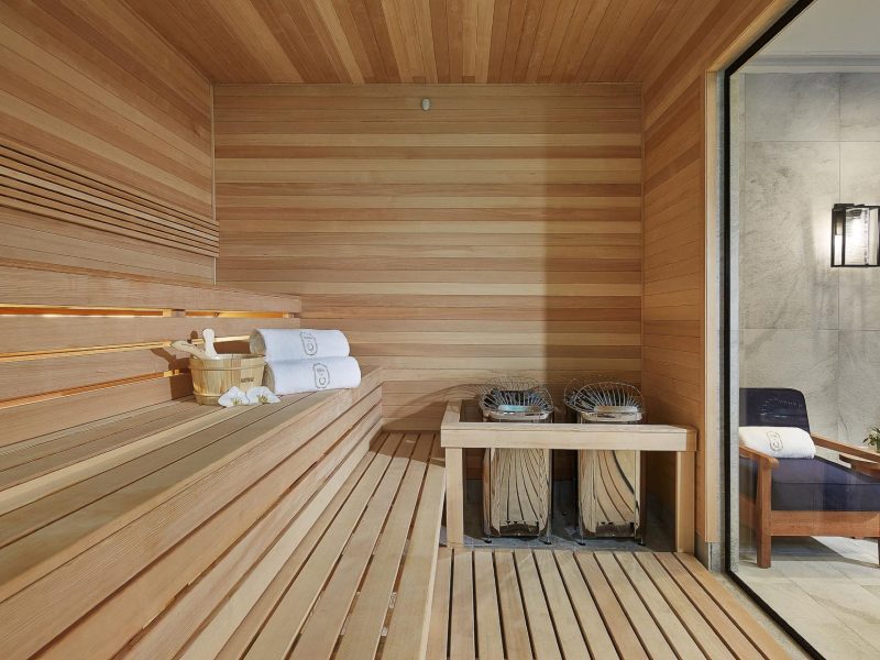 A wooden sauna room with tiered seating on the left, rolled white towels, and a small stand holding a bucket and ladle. Two heaters with rocks are on the right. The open door shows chairs and a small table outside the sauna. 124 on Queen Hotel and Spa | Hotel & Spa Niagara-on-the-lake | 1 (855) 988-4552