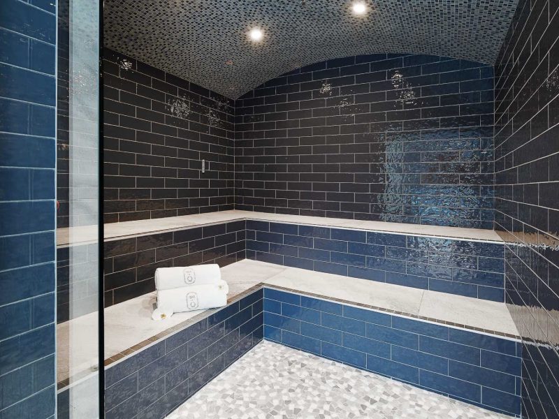 Luxurious steam room with glossy dark blue tiles and a mosaic tile floor. The ceiling features recessed lighting. Benches line the walls, each topped with white folded towels. 124 on Queen Hotel and Spa | Hotel & Spa Niagara-on-the-lake | 1 (855) 988-4552