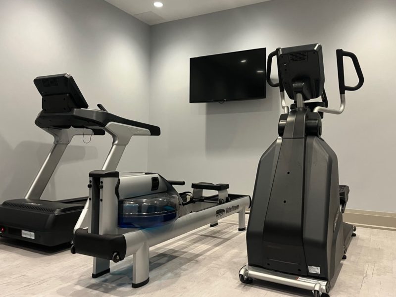 A gym room featuring a treadmill, a rowing machine with a blue water tank, and an elliptical trainer. A flat-screen TV is mounted on the wall above the equipment. The room has light gray walls and flooring. 124 on Queen Hotel and Spa | Hotel & Spa Niagara-on-the-lake | 1 (855) 988-4552
