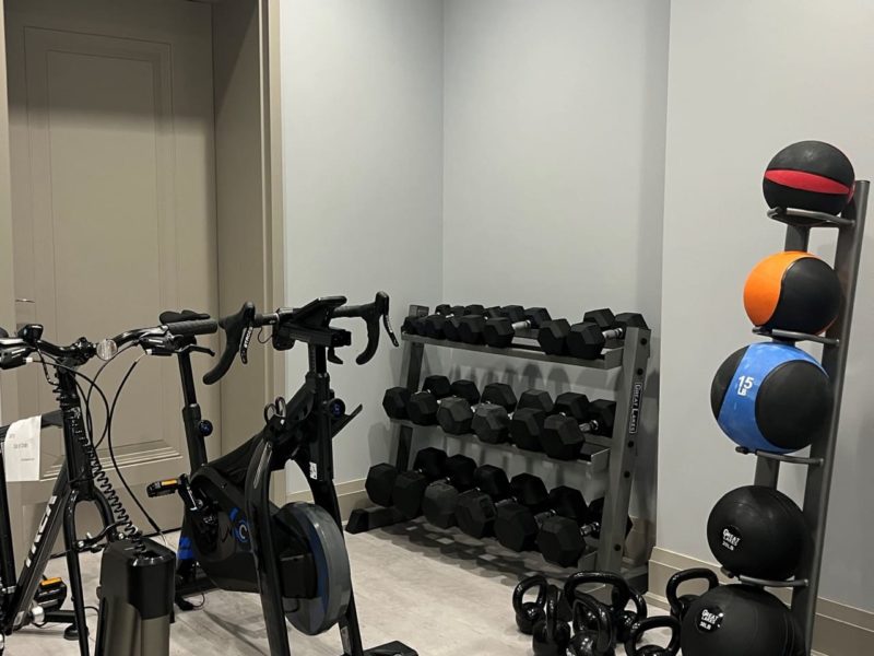 A small home gym with exercise bikes, a rack of dumbbells, kettlebells, medicine balls, and a step platform. The room has light gray walls and flooring, with recessed lighting above. 124 on Queen Hotel and Spa | Hotel & Spa Niagara-on-the-lake | 1 (855) 988-4552