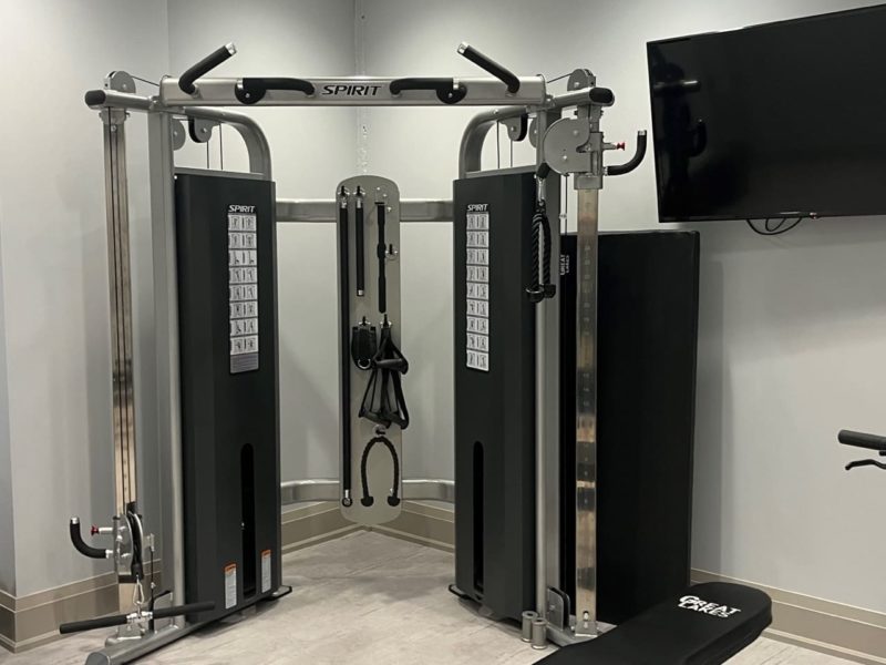 A modern gym room featuring a multifunctional cable machine with pulleys and attachments. A flat weight bench is positioned in the foreground. The room has light-colored walls and flooring, with a wall-mounted TV in the corner. 124 on Queen Hotel and Spa | Hotel & Spa Niagara-on-the-lake | 1 (855) 988-4552