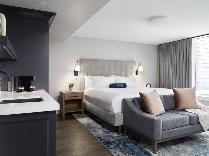A modern hotel room with a large bed, gray upholstered headboard, and dark throw pillows. A small couch sits at the foot of the bed. On the left, theres a kitchenette with a sink and coffee maker. The room has a large window with curtains. 124 on Queen Hotel and Spa | Hotel & Spa Niagara-on-the-lake | 1 (855) 988-4552