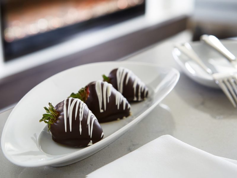 Three chocolate-covered strawberries with white chocolate drizzle are arranged on a white oval plate. The plate is on a table next to a folded napkin and cutlery in the background. 124 on Queen Hotel and Spa | Hotel & Spa Niagara-on-the-lake | 1 (855) 988-4552