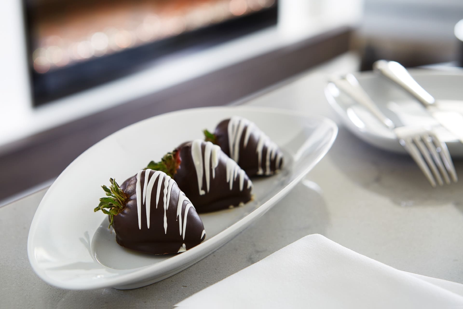 Three chocolate-covered strawberries with white chocolate drizzle are arranged on a white oval plate. The plate is on a table next to a folded napkin and cutlery in the background. 124 on Queen Hotel and Spa | Hotel & Spa Niagara-on-the-lake | 1 (855) 988-4552