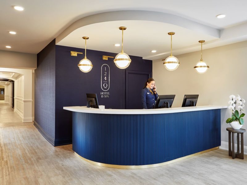 The image shows a hotel reception area with a curved desk in navy blue. A staff member in uniform is speaking on the phone. Behind her is a sign for 124 Hotel & Spa. Three globe pendant lights hang from the ceiling. A hallway extends to the left. 124 on Queen Hotel and Spa | Hotel & Spa Niagara-on-the-lake | 1 (855) 988-4552