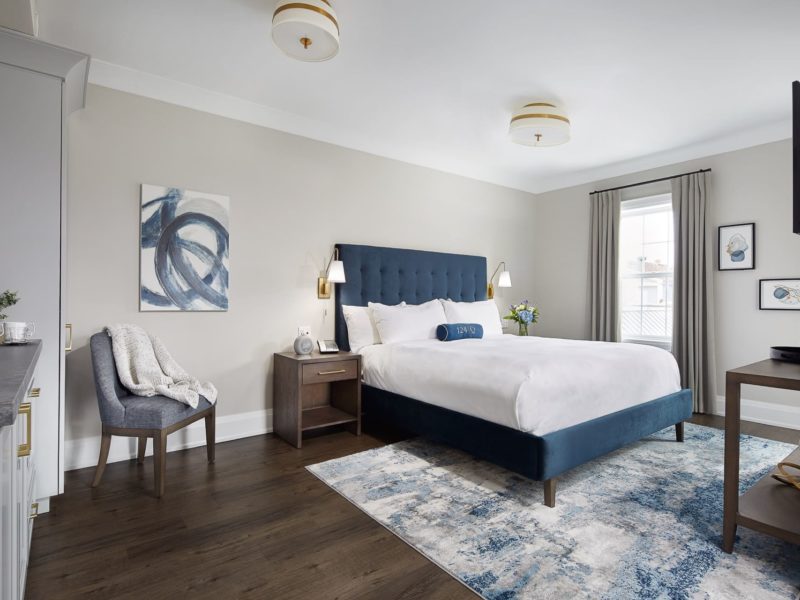 A modern bedroom with a dark blue upholstered bed, a blue and white abstract rug, and a white duvet. The room has a gray chair with a blanket, a wooden nightstand with a lamp, and a window with gray curtains. Wall art is hung above the chair. 124 on Queen Hotel and Spa | Hotel & Spa Niagara-on-the-lake | 1 (855) 988-4552