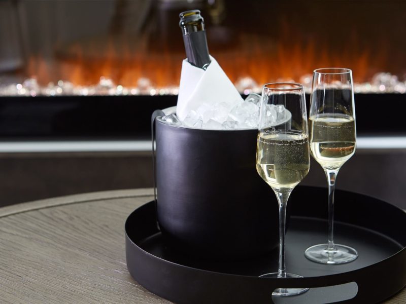 A bottle of champagne sits chilled in a black ice bucket on a round tray, accompanied by two filled champagne flutes. The scene is set on a wooden table in front of a warm, glowing fireplace. 124 on Queen Hotel and Spa | Hotel & Spa Niagara-on-the-lake | 1 (855) 988-4552