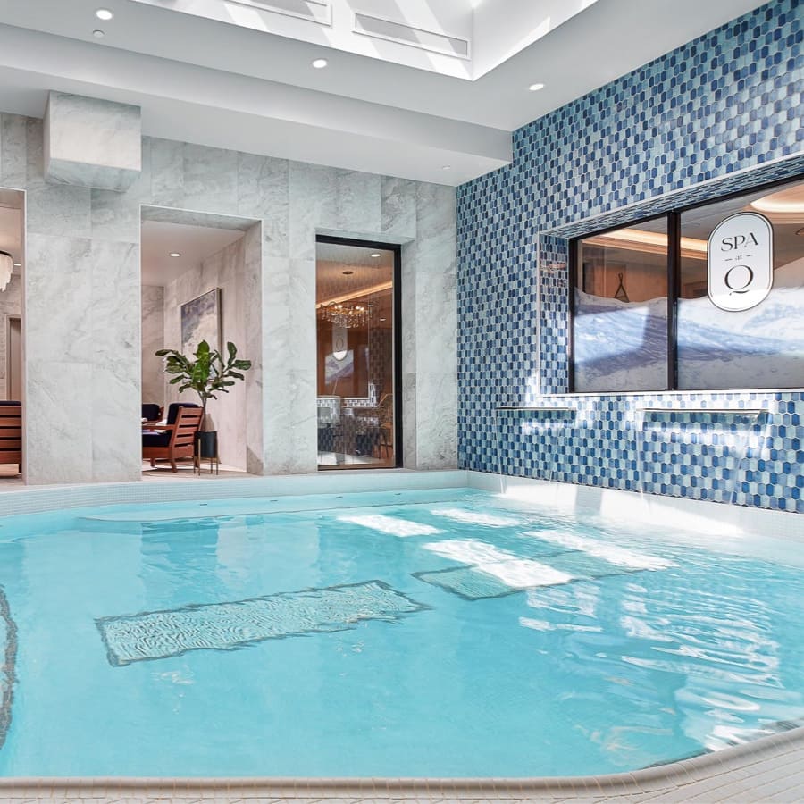 Indoor pool area with a small, clear turquoise pool featuring underwater benches. The walls have blue and white checkered tiles and large windows. A doorway leads to a room with seating and a plant, creating a luxurious and relaxing atmosphere. 124 on Queen Hotel and Spa | Hotel & Spa Niagara-on-the-lake | 1 (855) 988-4552