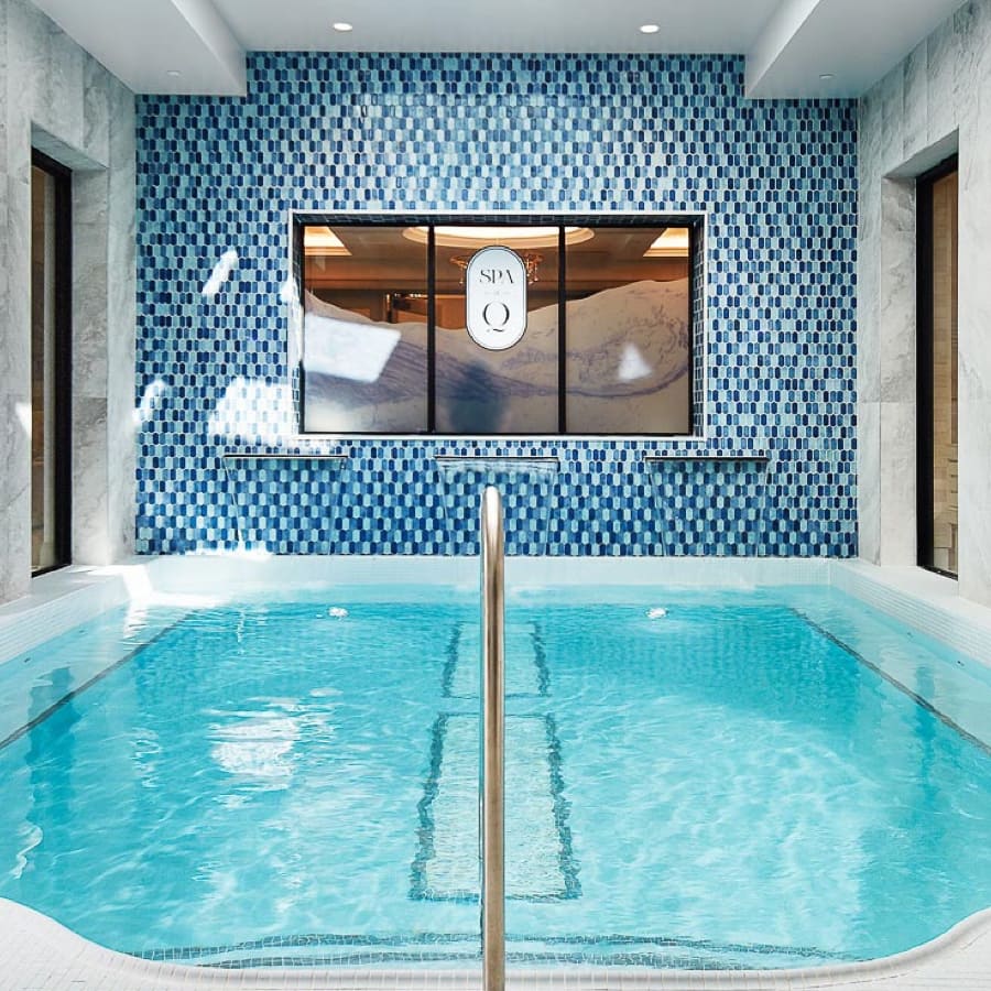 A small indoor pool with clear water is surrounded by white and blue mosaic tiles. A metal handrail leads into the pool. A window labeled SPA Q is centered on the far wall, adding a luxurious touch. 124 on Queen Hotel and Spa | Hotel & Spa Niagara-on-the-lake | 1 (855) 988-4552