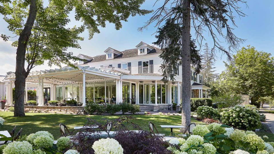The Gate House Wedding Venue is a historic building surrounded by lush greenery, featuring a white pergola adorned with string lights. Tall trees and blooming hydrangeas grace the foreground, while large windows and a classic roof with dormers enhance its charm under a clear sky. 124 on Queen Hotel and Spa | Hotel & Spa Niagara-on-the-lake | 1 (855) 988-4552