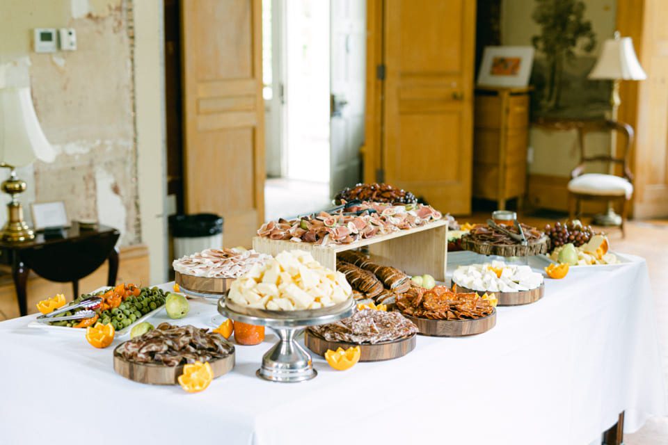 A large charcuterie table is elegantly set up with a variety of meats, cheeses, nuts, and fruits, including grapes and orange slices. The table is covered with a white cloth and surrounded by softly lit, rustic decor in a spacious room. 124 on Queen Hotel and Spa | Hotel & Spa Niagara-on-the-lake | 1 (855) 988-4552