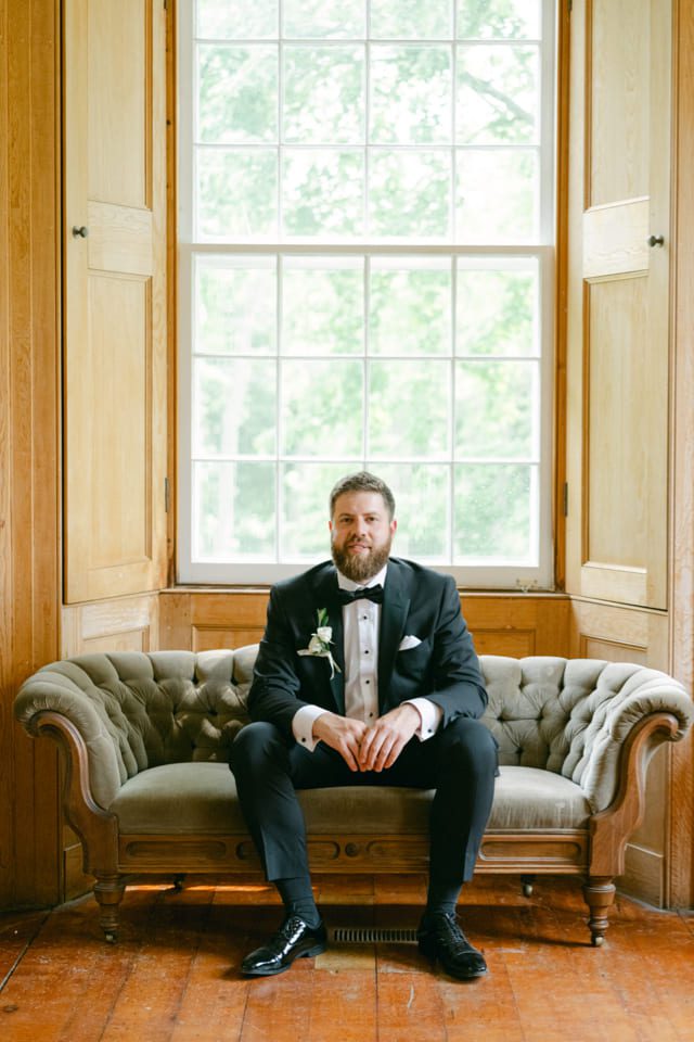 Man in a tuxedo sits on a vintage sofa in front of a large window. The room has wooden paneling. He has a boutonniere on his lapel and is looking at the camera. Natural light illuminates the scene. 124 on Queen Hotel and Spa | Hotel & Spa Niagara-on-the-lake | 1 (855) 988-4552