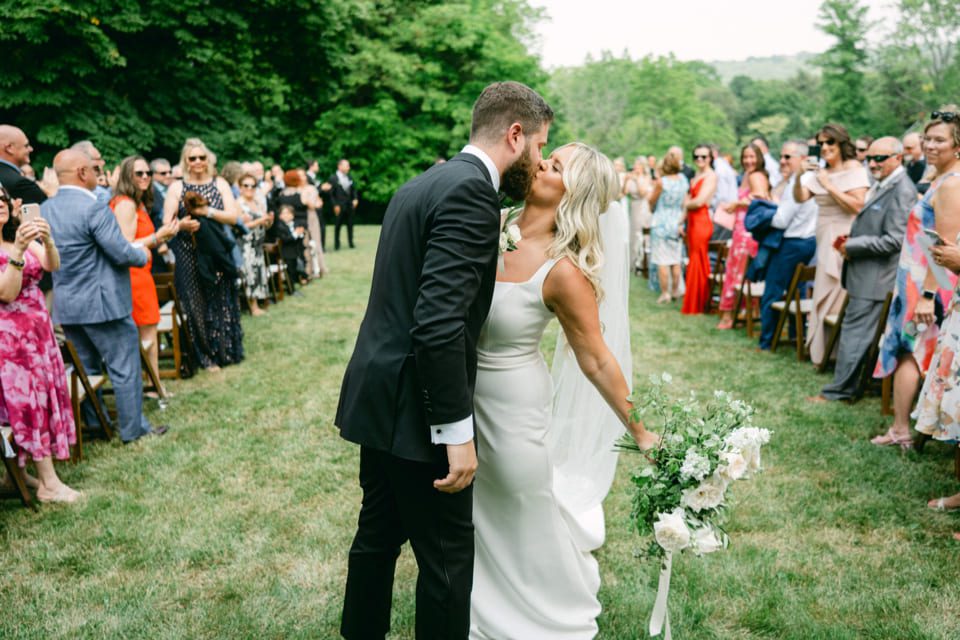 A bride and groom share a kiss in an outdoor wedding ceremony. The bride, in a white dress, holds a bouquet. The groom wears a black suit. Guests surround them, clapping and taking photos. Trees and greenery form the backdrop. 124 on Queen Hotel and Spa | Hotel & Spa Niagara-on-the-lake | 1 (855) 988-4552