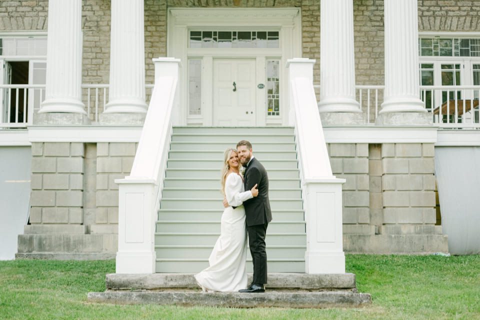 A couple stands on the steps of a grand building, embracing and smiling. The bride is in a long white dress, and the groom is in a black suit. The building features large white columns and a stone facade. 124 on Queen Hotel and Spa | Hotel & Spa Niagara-on-the-lake | 1 (855) 988-4552