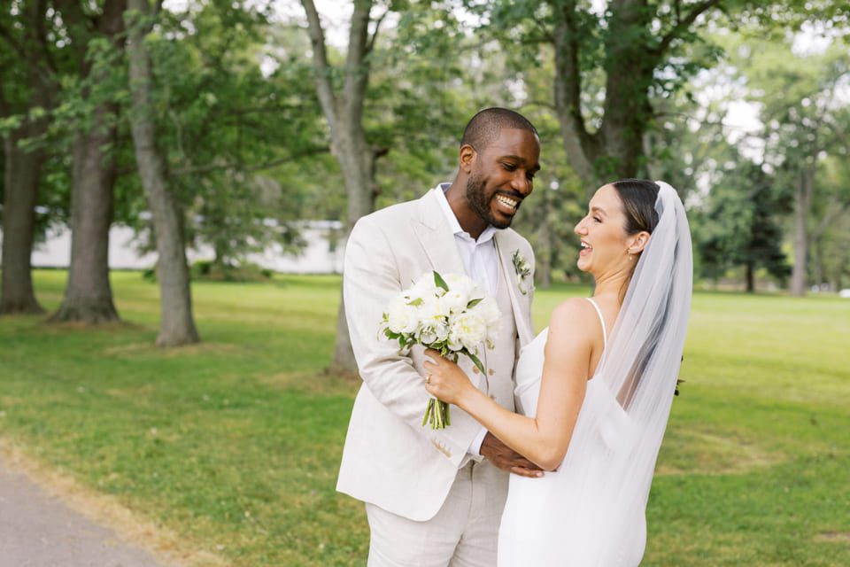 A happy couple smiling at each other in a lush, green park setting. The groom is wearing a light suit, and the bride is in a white dress and veil, holding a bouquet of white flowers. Trees and a grassy area are in the background. 124 on Queen Hotel and Spa | Hotel & Spa Niagara-on-the-lake | 1 (855) 988-4552