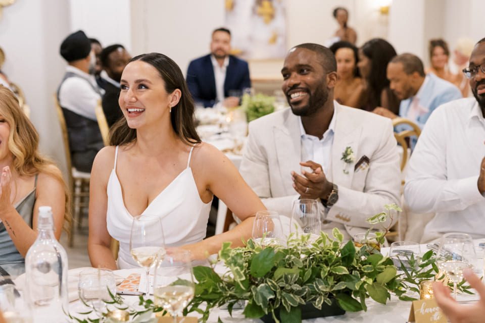 A smiling woman in a white dress sits beside a man in a beige suit at a formal event. They are at a table decorated with greenery and glassware. Other guests are seated around them, and the atmosphere is joyful and elegant. 124 on Queen Hotel and Spa | Hotel & Spa Niagara-on-the-lake | 1 (855) 988-4552