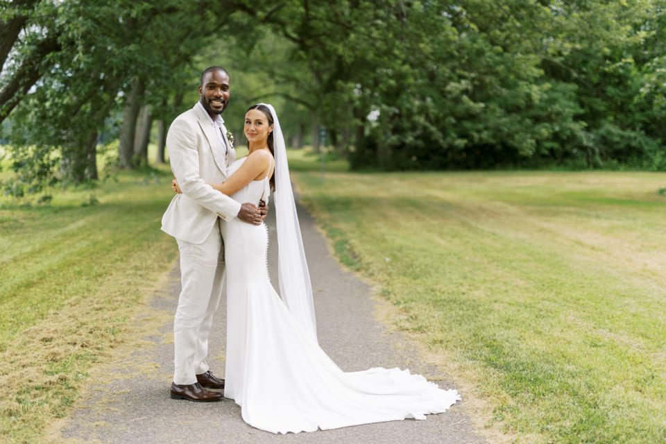 A bride and groom stand together on a paved path surrounded by grass and trees. The groom wears a light suit, and the bride wears a long white dress with a veil. Both are smiling as they embrace, with greenery in the background. 124 on Queen Hotel and Spa | Hotel & Spa Niagara-on-the-lake | 1 (855) 988-4552