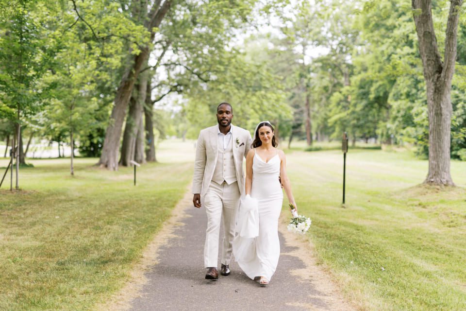 A bride and groom walk down a tree-lined path. The groom wears a light tan suit, and the bride wears a white dress, holding a bouquet. They appear happy and relaxed on a sunny day, surrounded by lush greenery. 124 on Queen Hotel and Spa | Hotel & Spa Niagara-on-the-lake | 1 (855) 988-4552