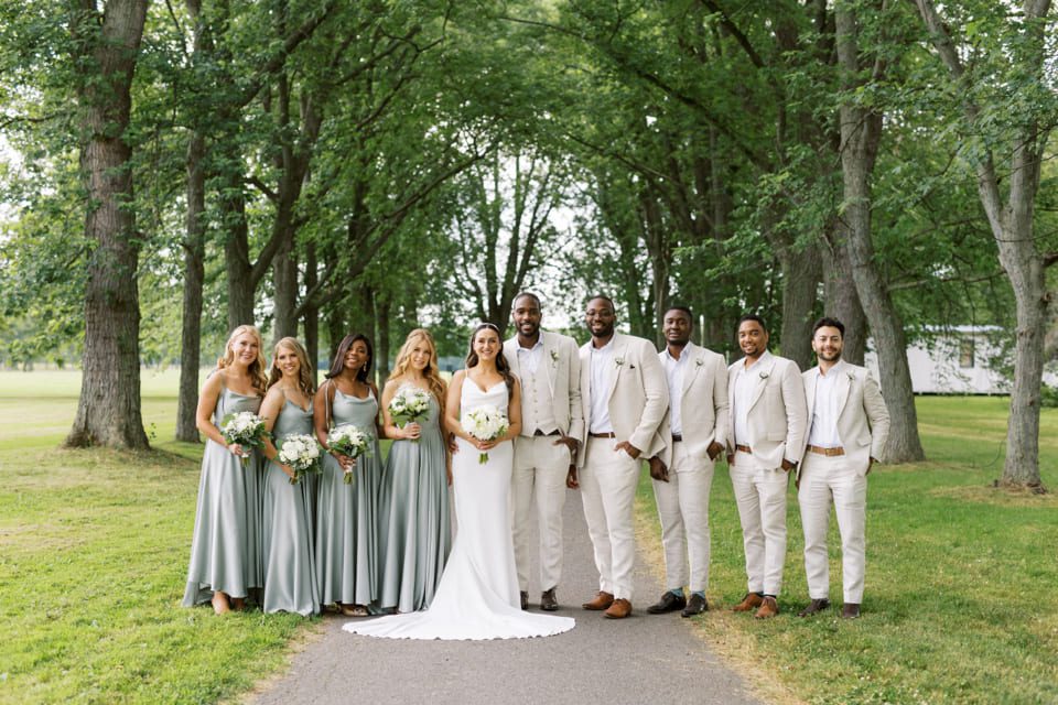 A wedding party stands on a tree-lined path. The bride is in a white gown, flanked by bridesmaids in silver dresses and groomsmen in light suits. Everyone is smiling and holding bouquets. The background features lush greenery and trees. 124 on Queen Hotel and Spa | Hotel & Spa Niagara-on-the-lake | 1 (855) 988-4552