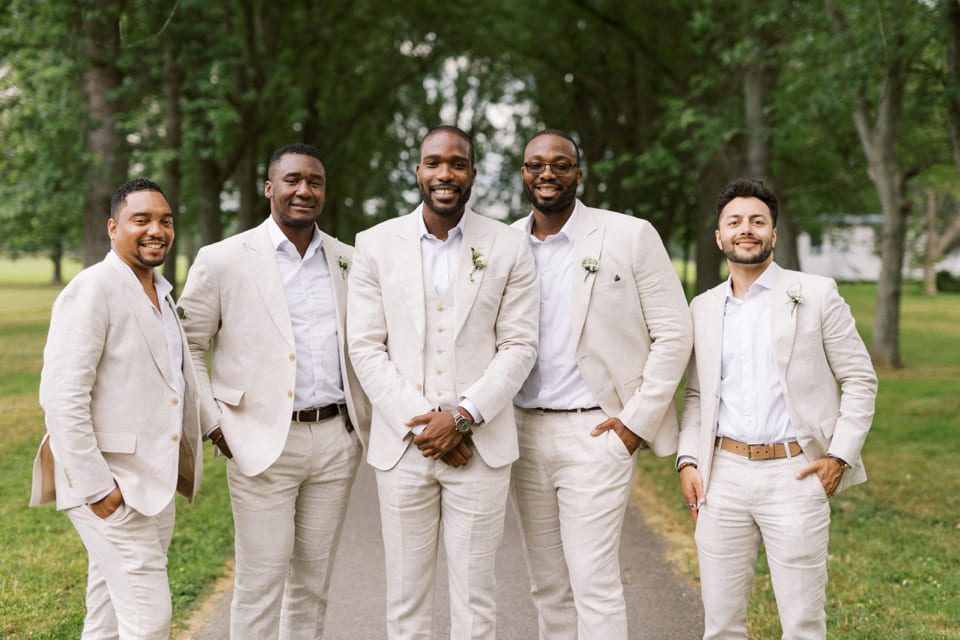 Five men in light beige suits stand together on a tree-lined path. They are smiling and appear to be at a formal outdoor event. Small white flowers are pinned on their lapels. The background features lush green trees and grass. 124 on Queen Hotel and Spa | Hotel & Spa Niagara-on-the-lake | 1 (855) 988-4552