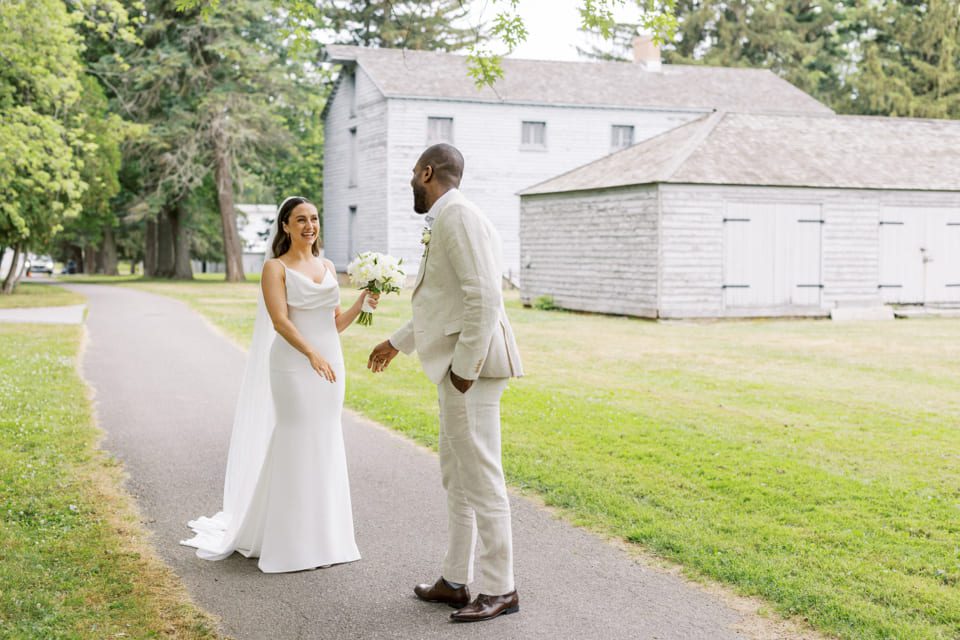 A bride in a white gown and veil holds a bouquet, smiling at a groom in a light beige suit. They stand on a path in front of a rustic building surrounded by greenery. 124 on Queen Hotel and Spa | Hotel & Spa Niagara-on-the-lake | 1 (855) 988-4552