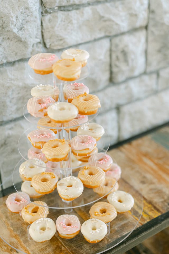 A tiered stand displaying various glazed mini donuts, some with white, pink, or cream-colored icing and sprinkles. The background features a stone wall and a wooden surface. 124 on Queen Hotel and Spa | Hotel & Spa Niagara-on-the-lake | 1 (855) 988-4552