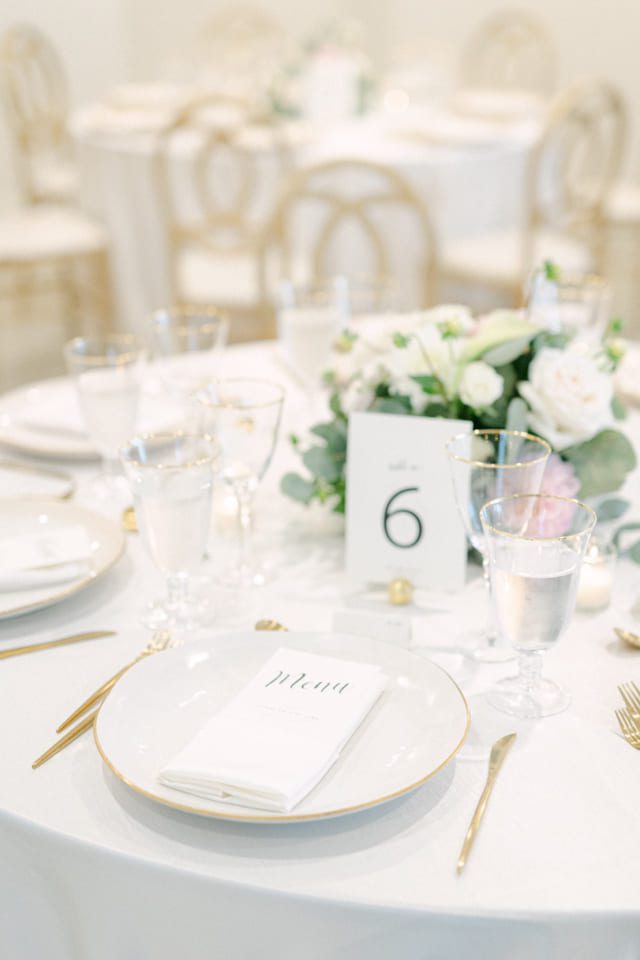 Elegant table setting with white plates, gold-rimmed glasses, gold cutlery, and white napkins. A table number 6 and a floral centerpiece with pastel flowers are in the center. Soft, warm lighting enhances the sophisticated ambiance. 124 on Queen Hotel and Spa | Hotel & Spa Niagara-on-the-lake | 1 (855) 988-4552